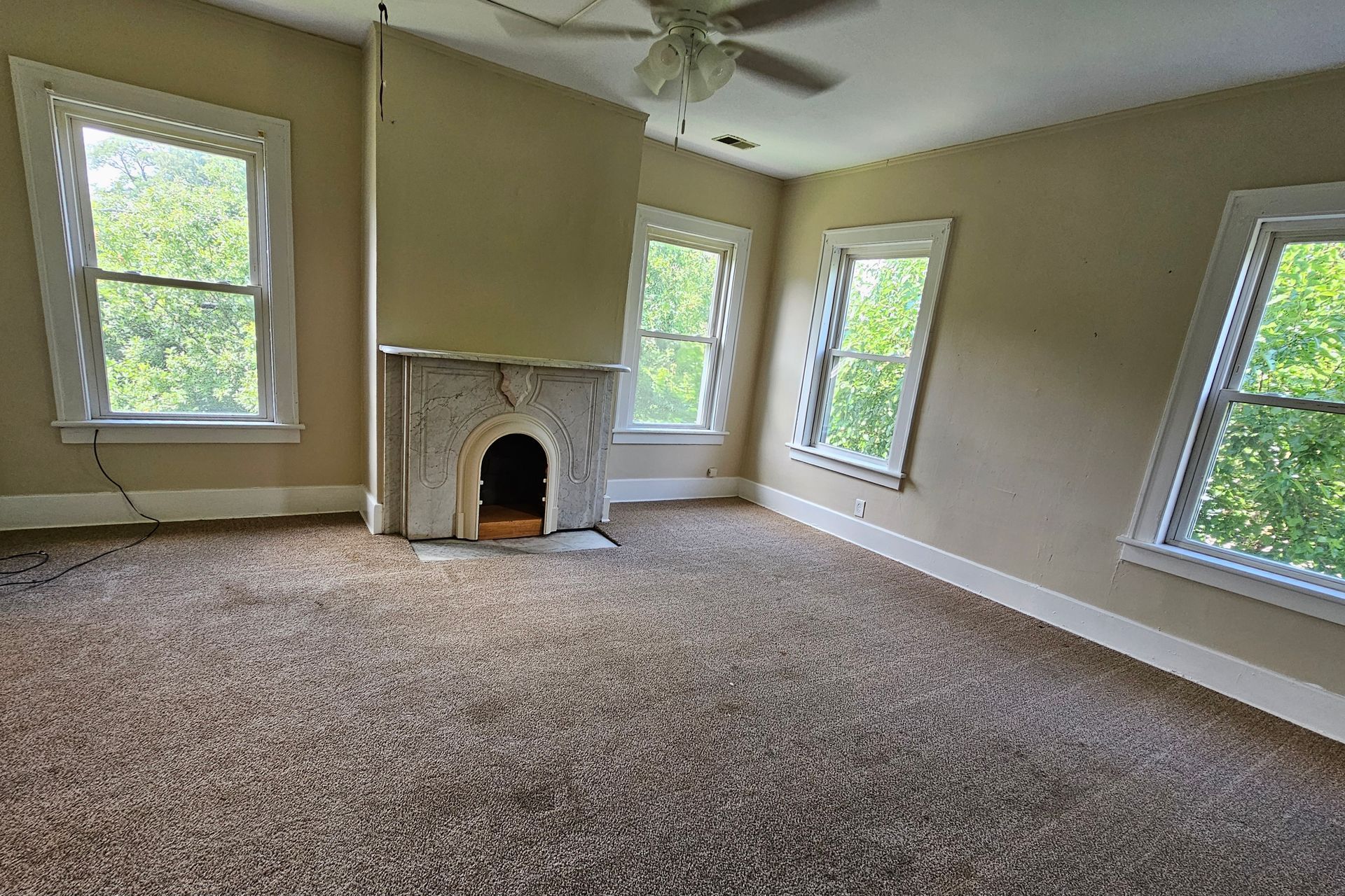 Empty beige living room with carpet, fireplace, ceiling fan, and large windows