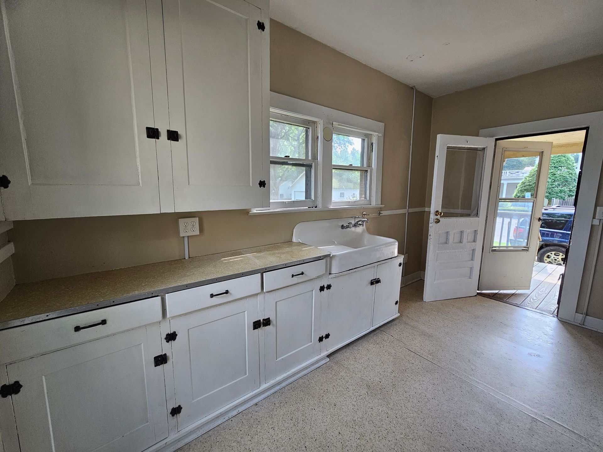 Bright kitchen with white cabinets, beige countertops, window over sink, and open door to driveway.