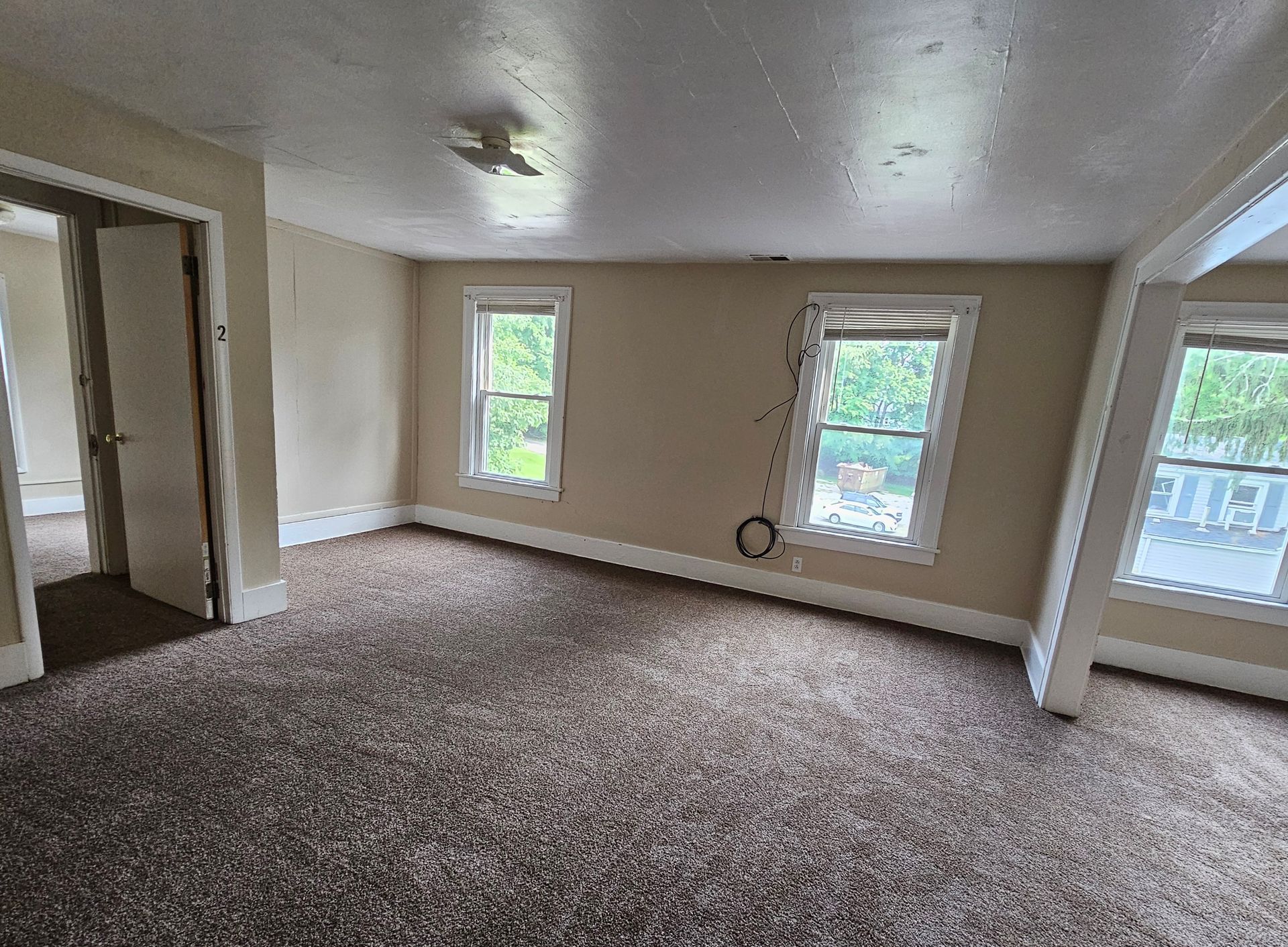 Empty beige living room with brown carpet, three windows, and a doorway to the hall