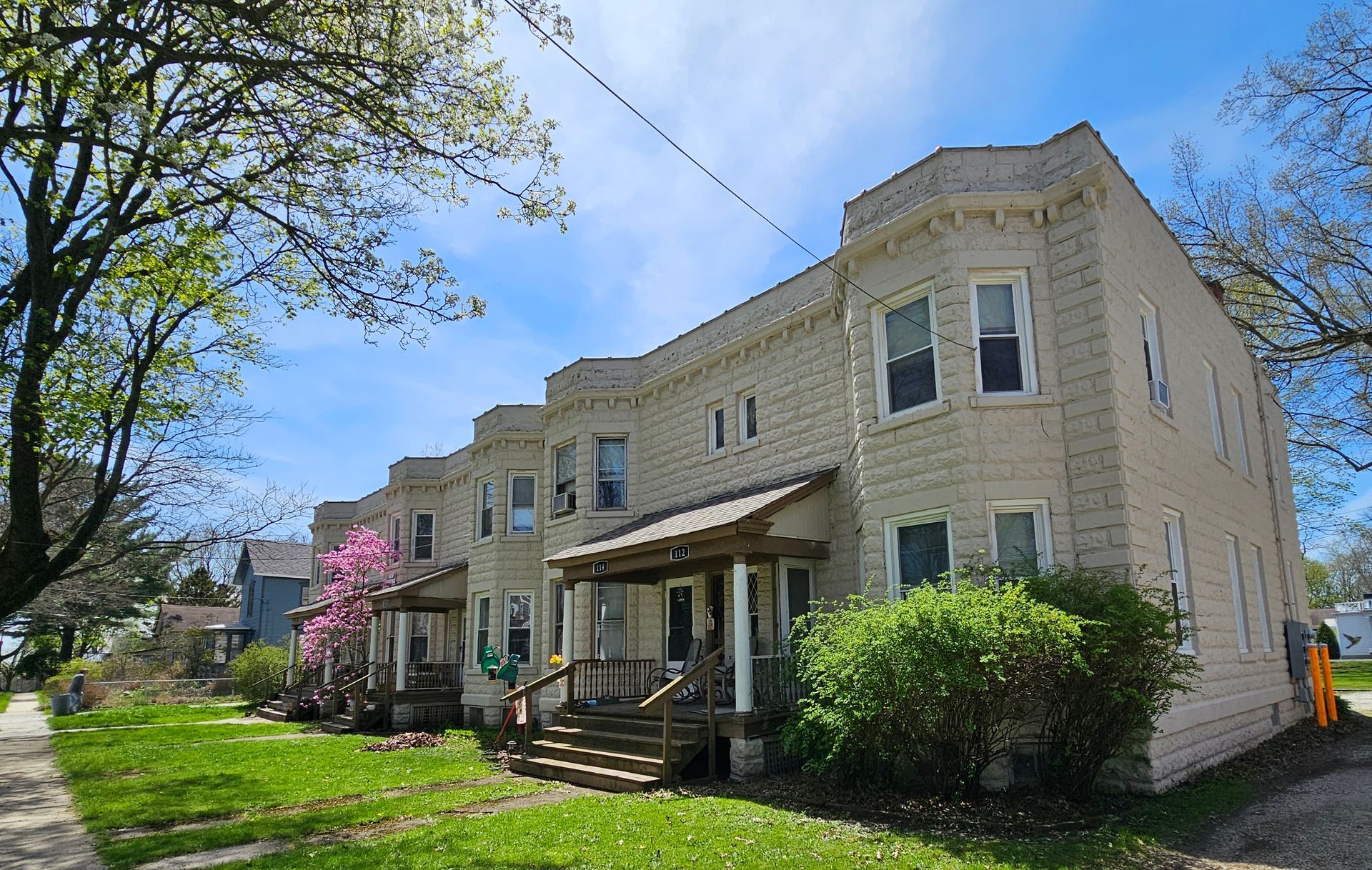 Row of beige townhouses with porches on a sunny street, trees and green lawn in front