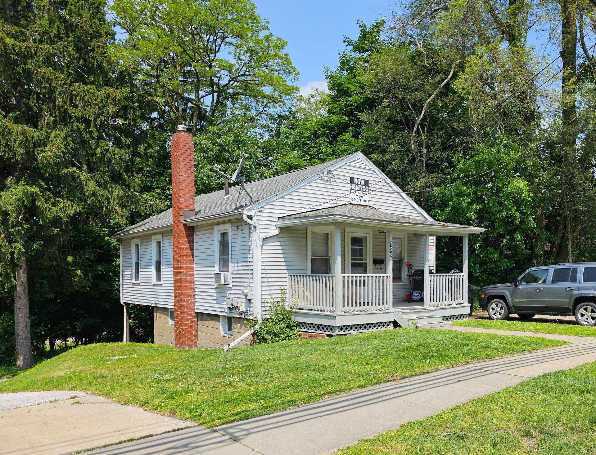 Small white house with porch, gray roof, brick chimney, and a parked SUV on a sunny tree-lined street