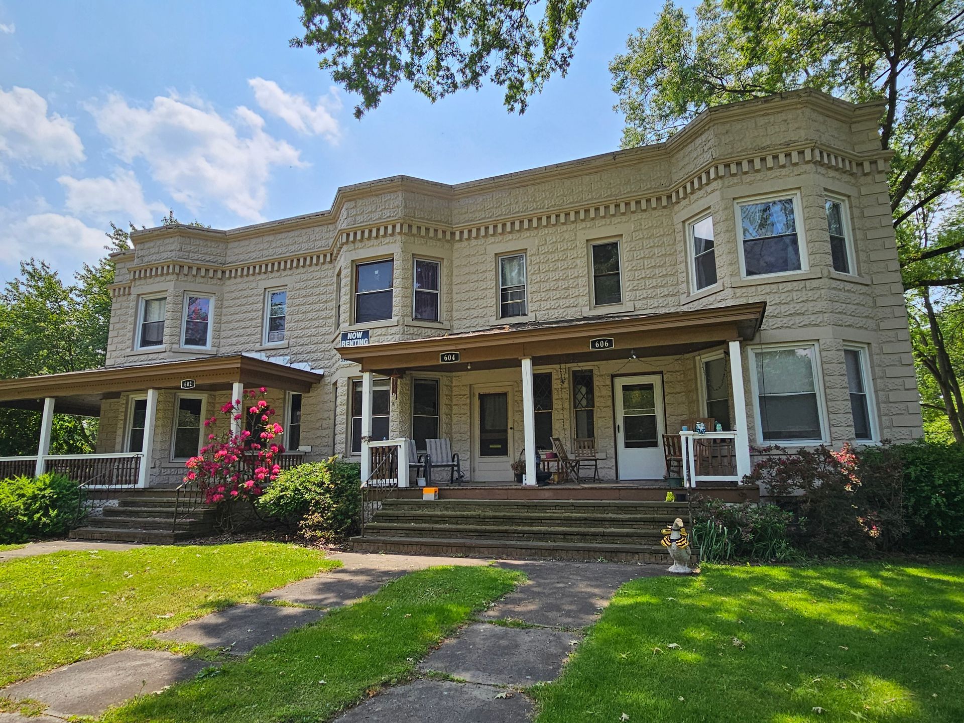 Two-story beige Victorian house with front porch, stone steps, and green lawn under a blue sky