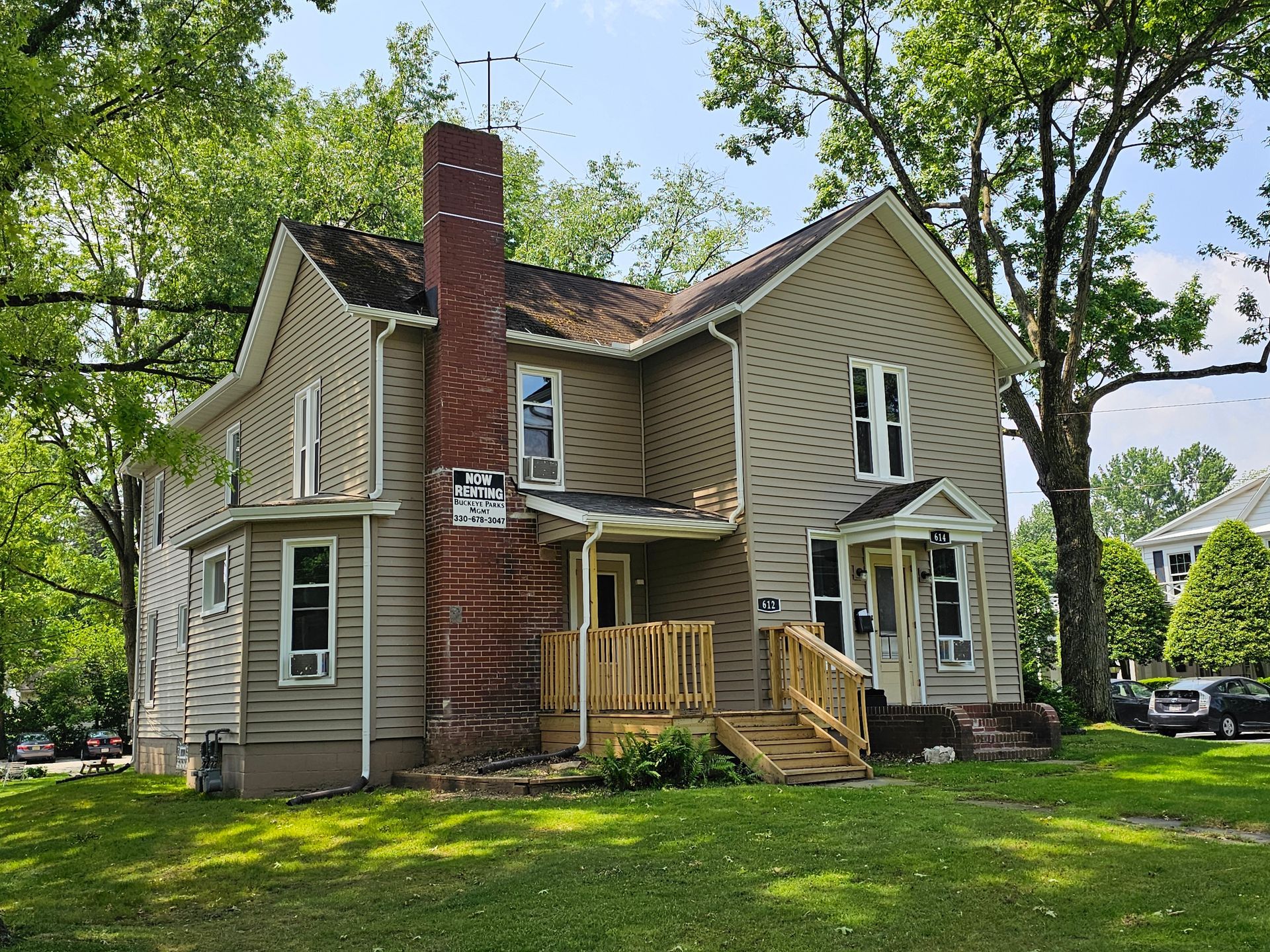 Old beige house with a red brick chimney, white trim, and a small wooden porch in a leafy yard
