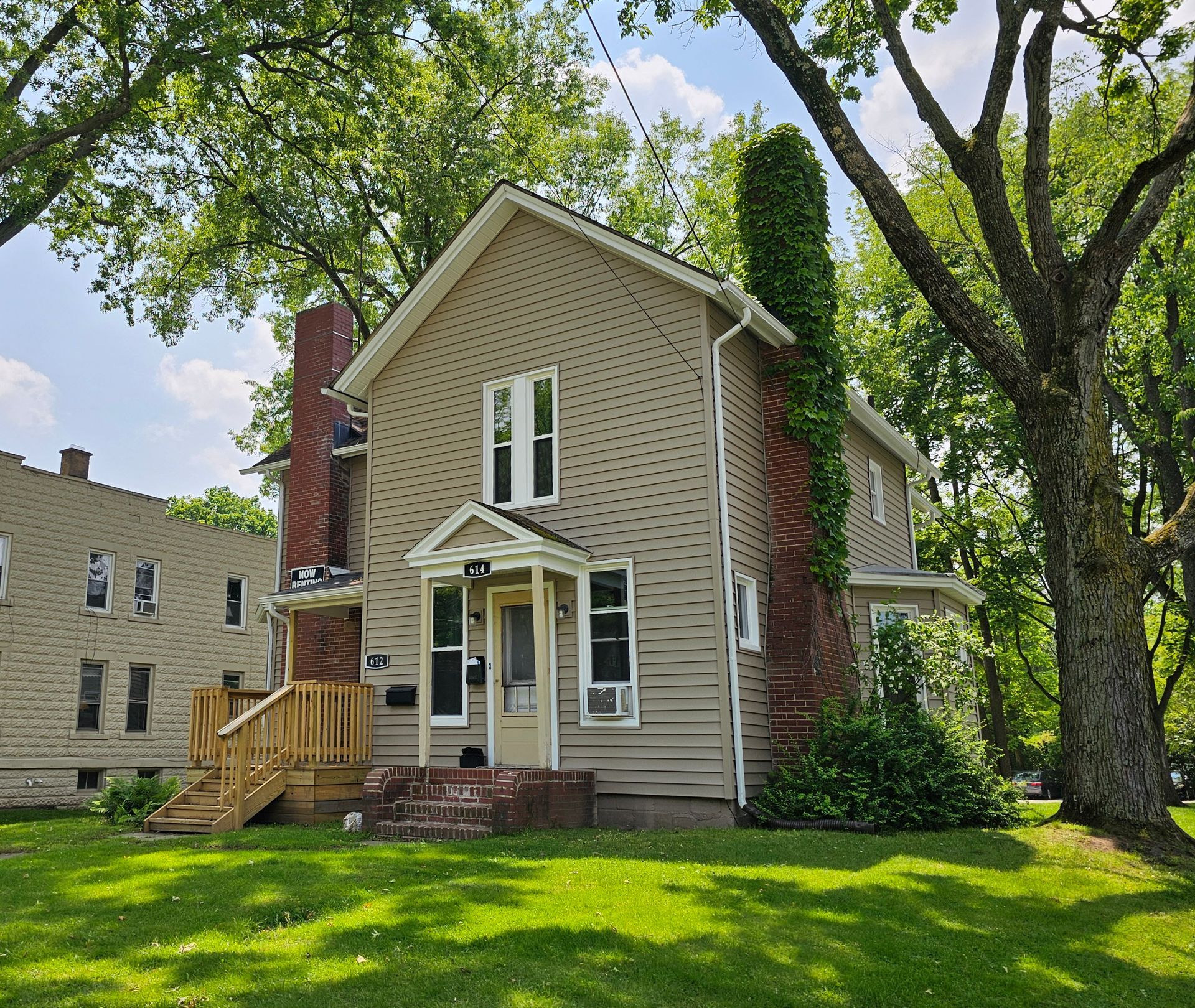 Beige two-story house with white trim, brick chimney, and large green lawn under trees.