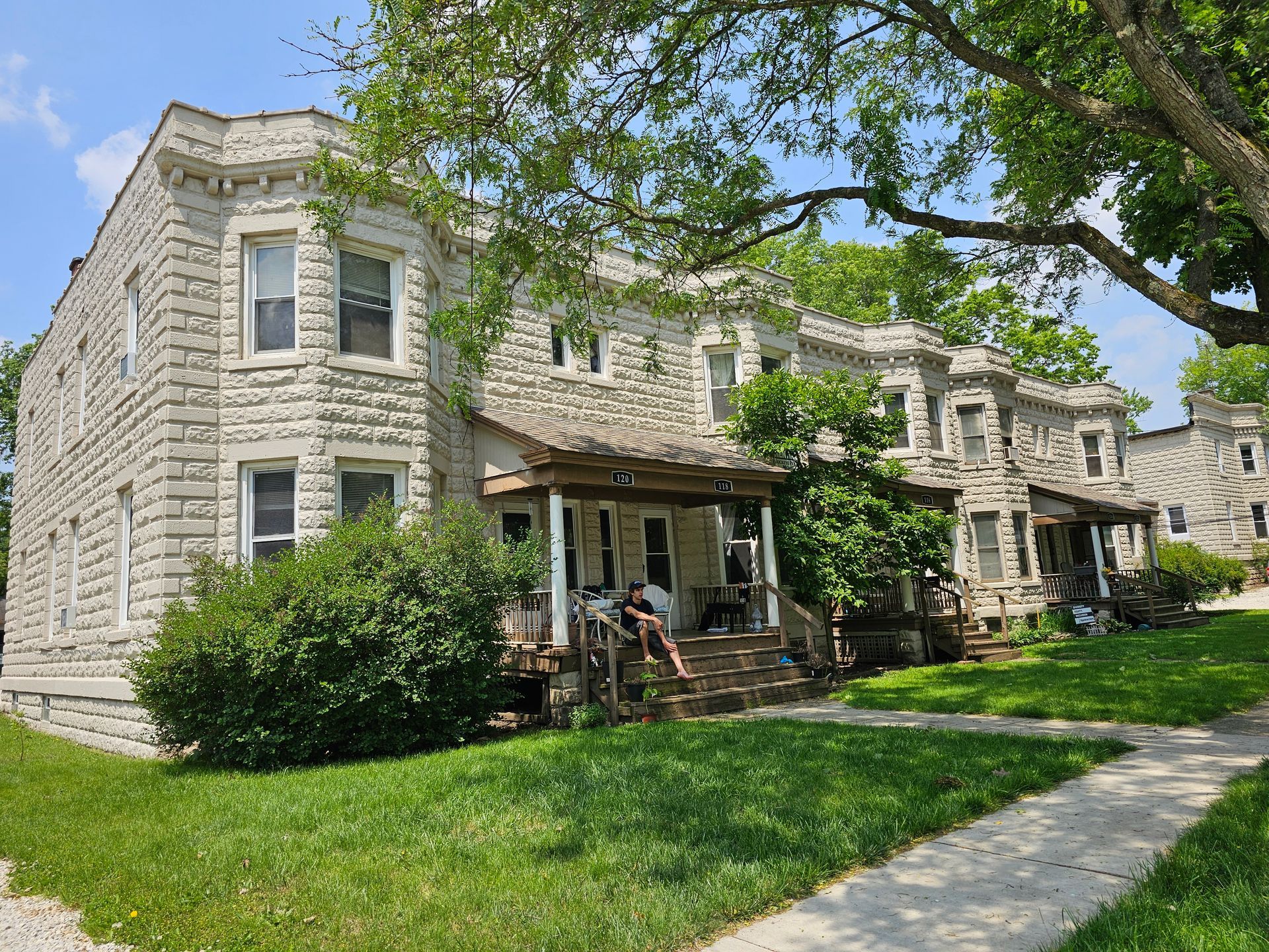 Stone apartment buildings with front porches, green lawn, and sidewalk under a leafy tree on a sunny day