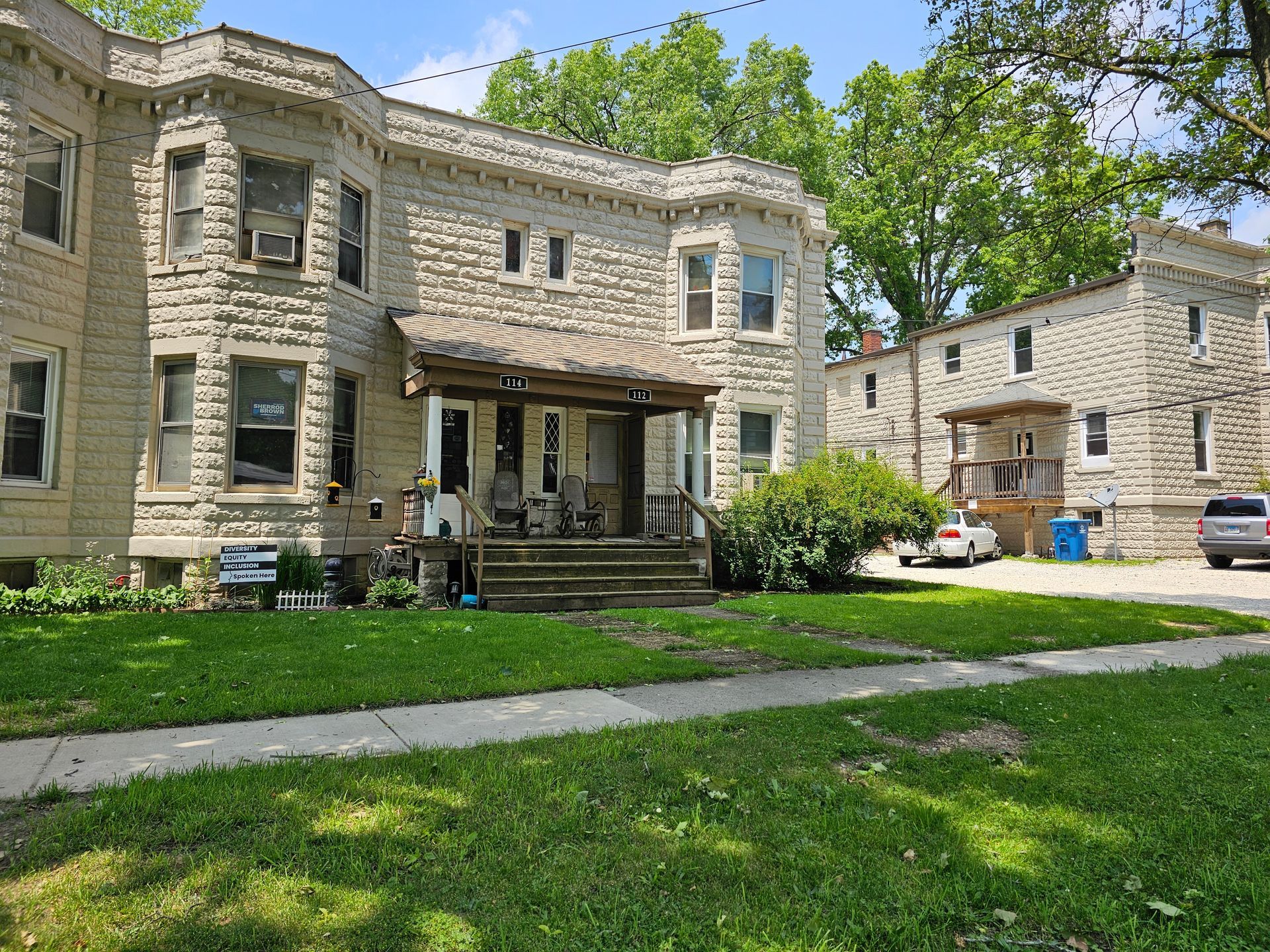 Stone apartment buildings with a grassy courtyard, sidewalk, and parked cars on a sunny day