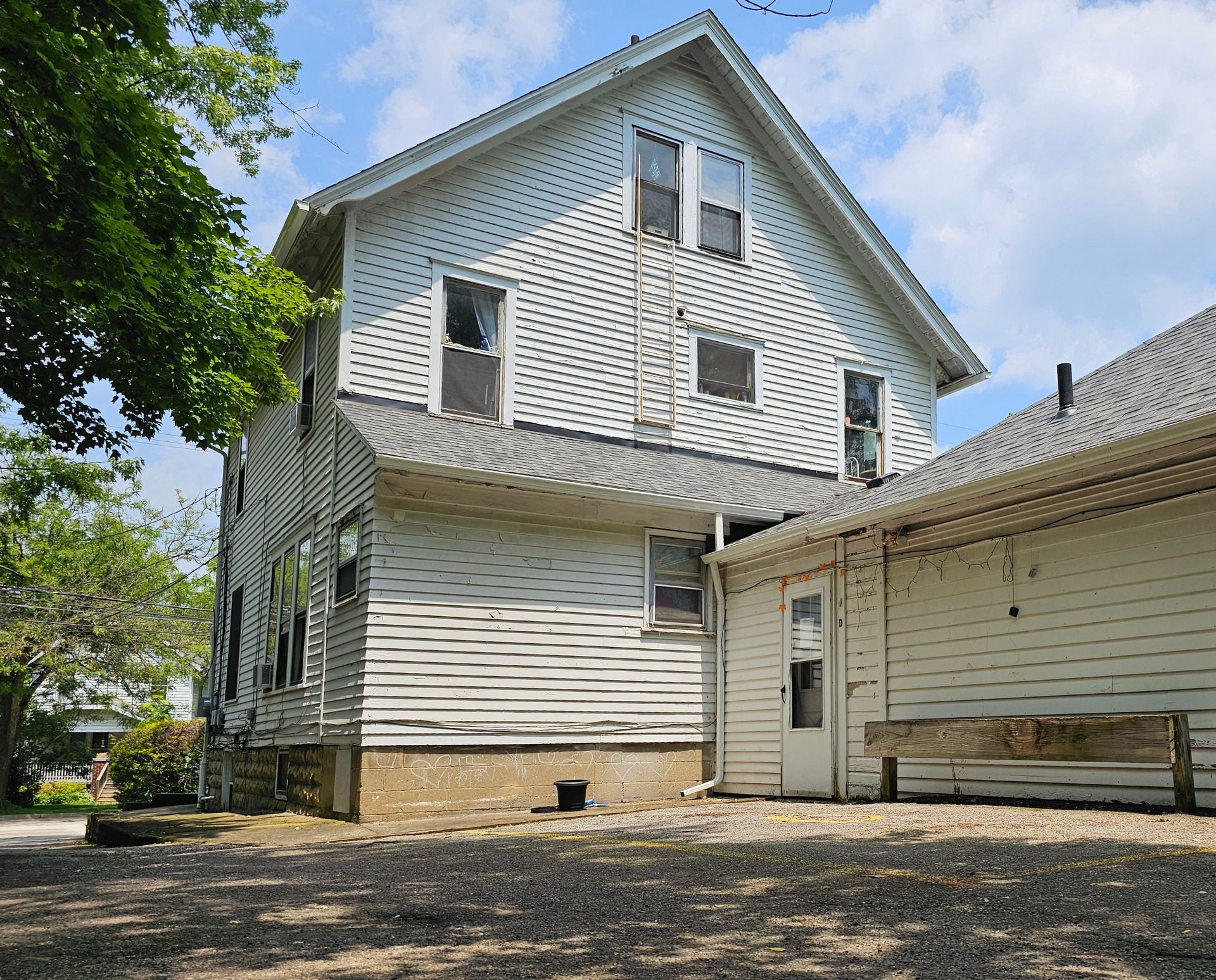 White two-story house with siding, side door, and gravel yard under a partly cloudy sky.
