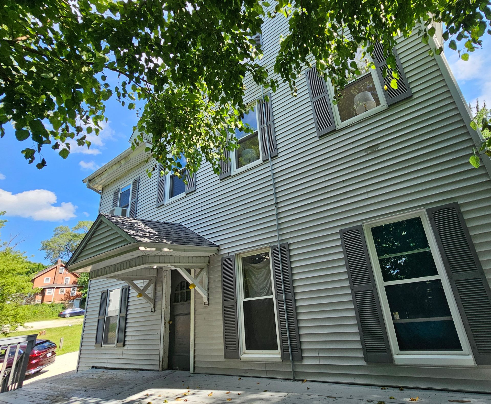 Gray house exterior with white trim, front awning, and large windows under leafy tree branches