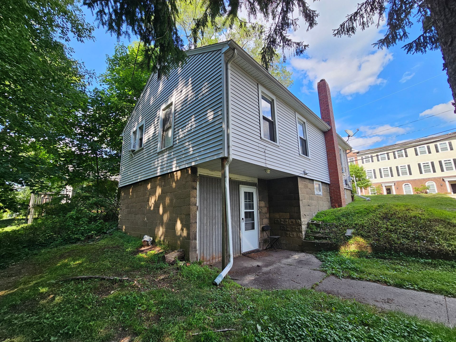 Small gray house with white trim and brick chimney, surrounded by trees and bushes under a blue sky