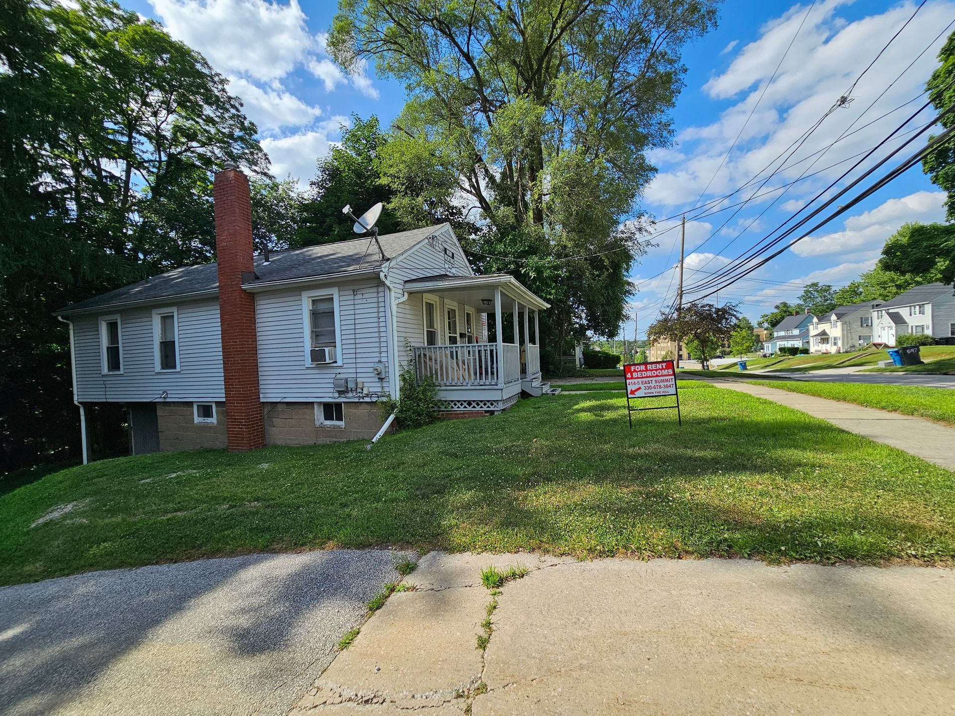 White house with a red brick chimney and porch on a grassy corner lot under a blue sky.