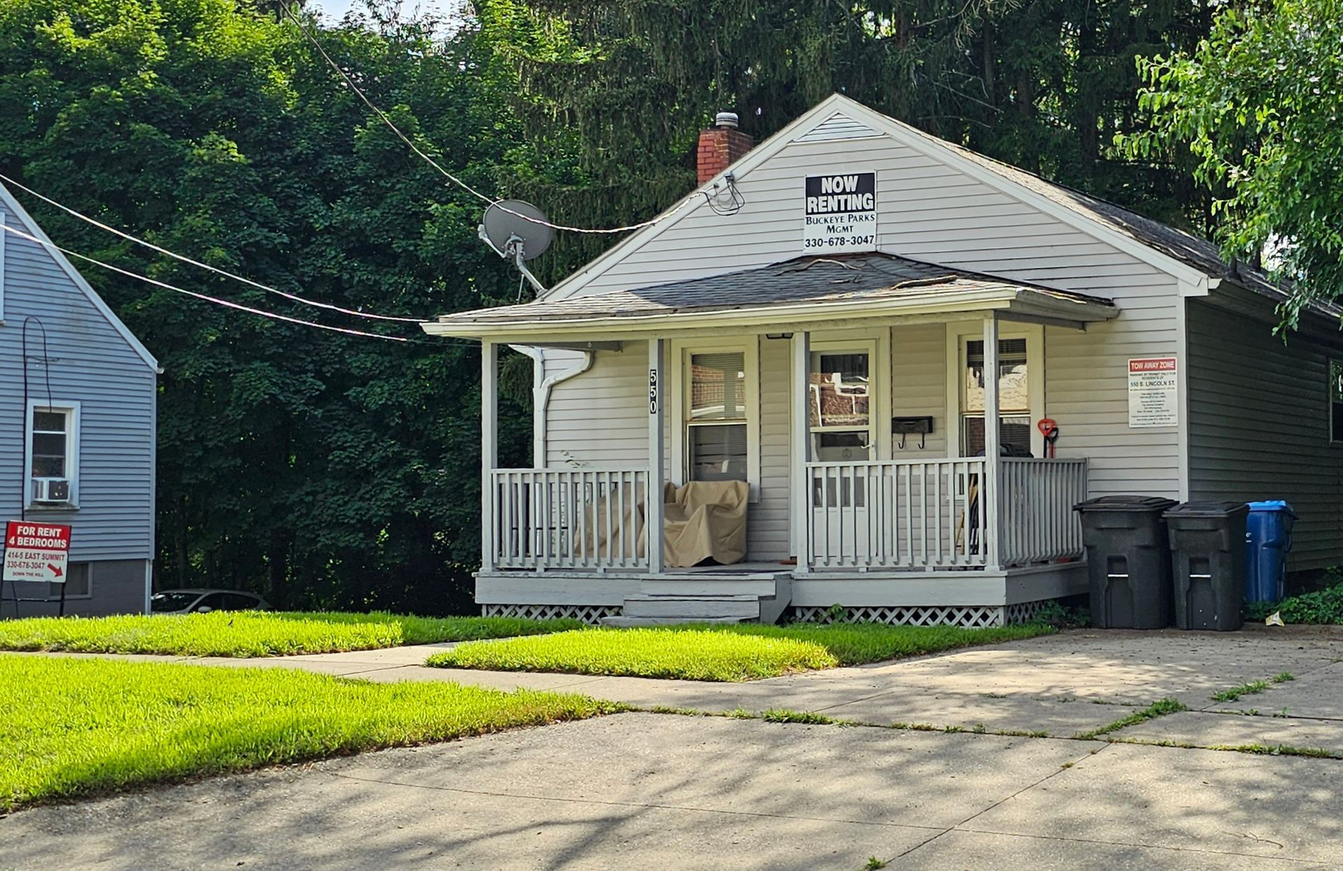 Small white house with a front porch, gray roof, and trash bins beside a driveway under trees.