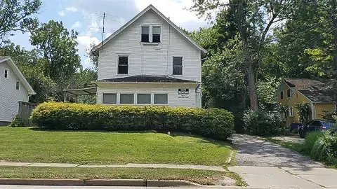White two-story house with green hedge, lawn, driveway, and trees on a sunny suburban street.