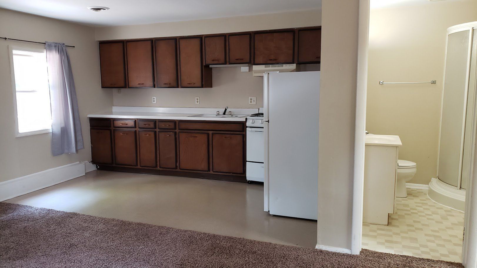 Empty apartment kitchen with wood cabinets, white fridge, beige carpet, and a small bathroom doorway