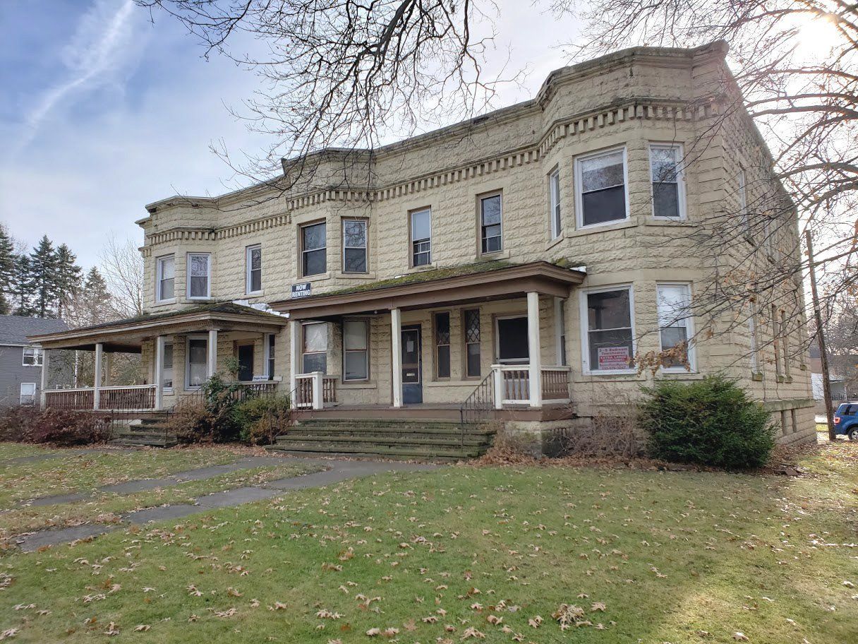 Historic two-story stone house with a porch and winter lawn, viewed from the front.