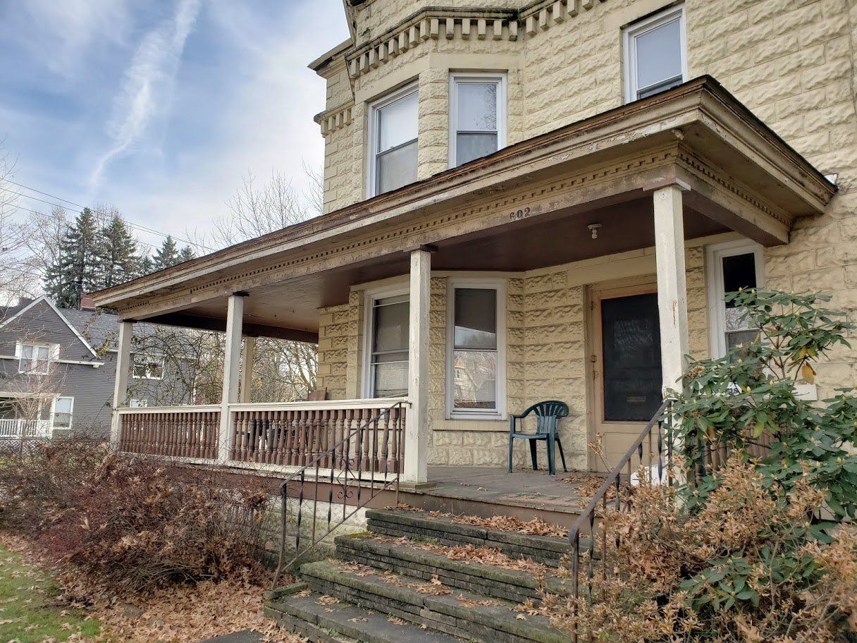 Front porch of a beige Victorian house with steps, railing, and a dark front door