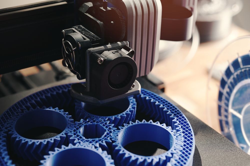 A 3D printer head printing a blue gear.