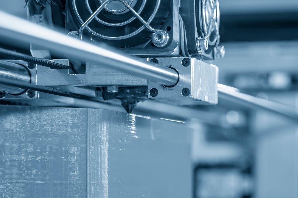 3D printer nozzle extruding filament onto a growing rectangular object, with a fan in the background.