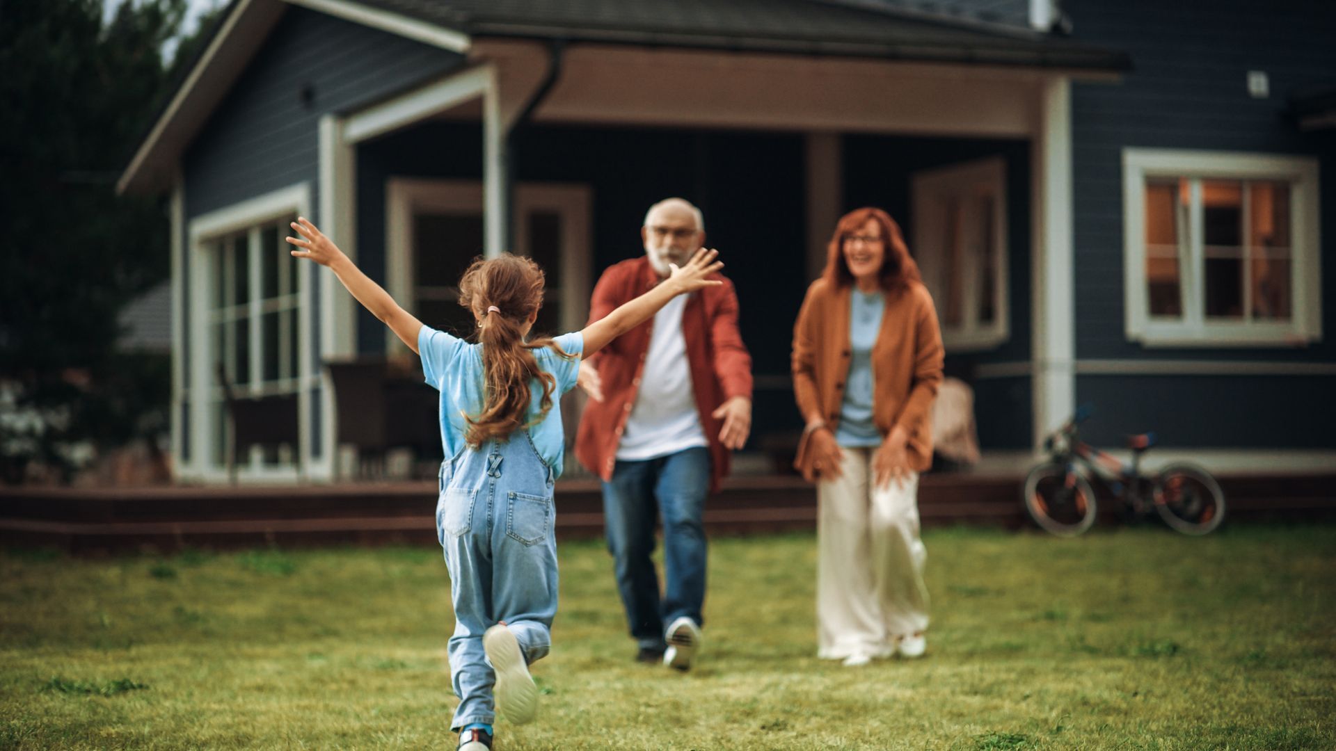 A little girl is running in front of a house with her grandparents.