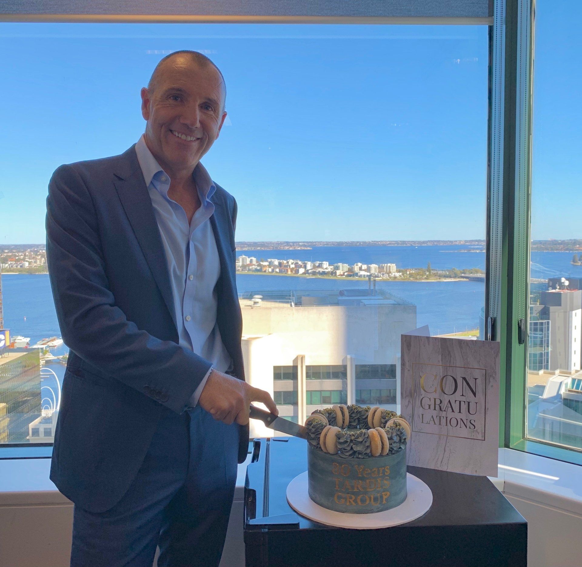 A man in a suit is cutting a cake in front of a window.
