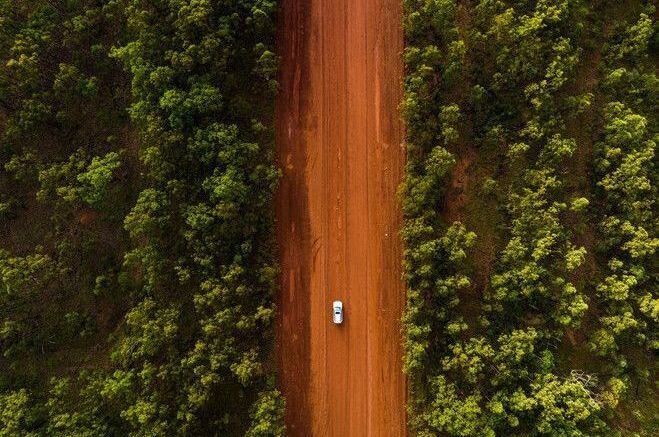 Aerial View of a Solitary White Car Driving on a Reddish Dirt Road Flanked by Lush Green Trees — NT Driver Guide - Escorted Touring & Transfers in Darwin City, NT