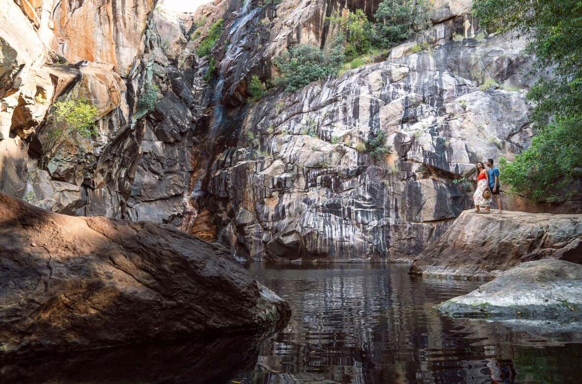 A Couple Stands by a Serene Pool Surrounded by Rugged Cliffs and Cascading Water — NT Driver Guide - Escorted Touring & Transfers in Darwin City, NT