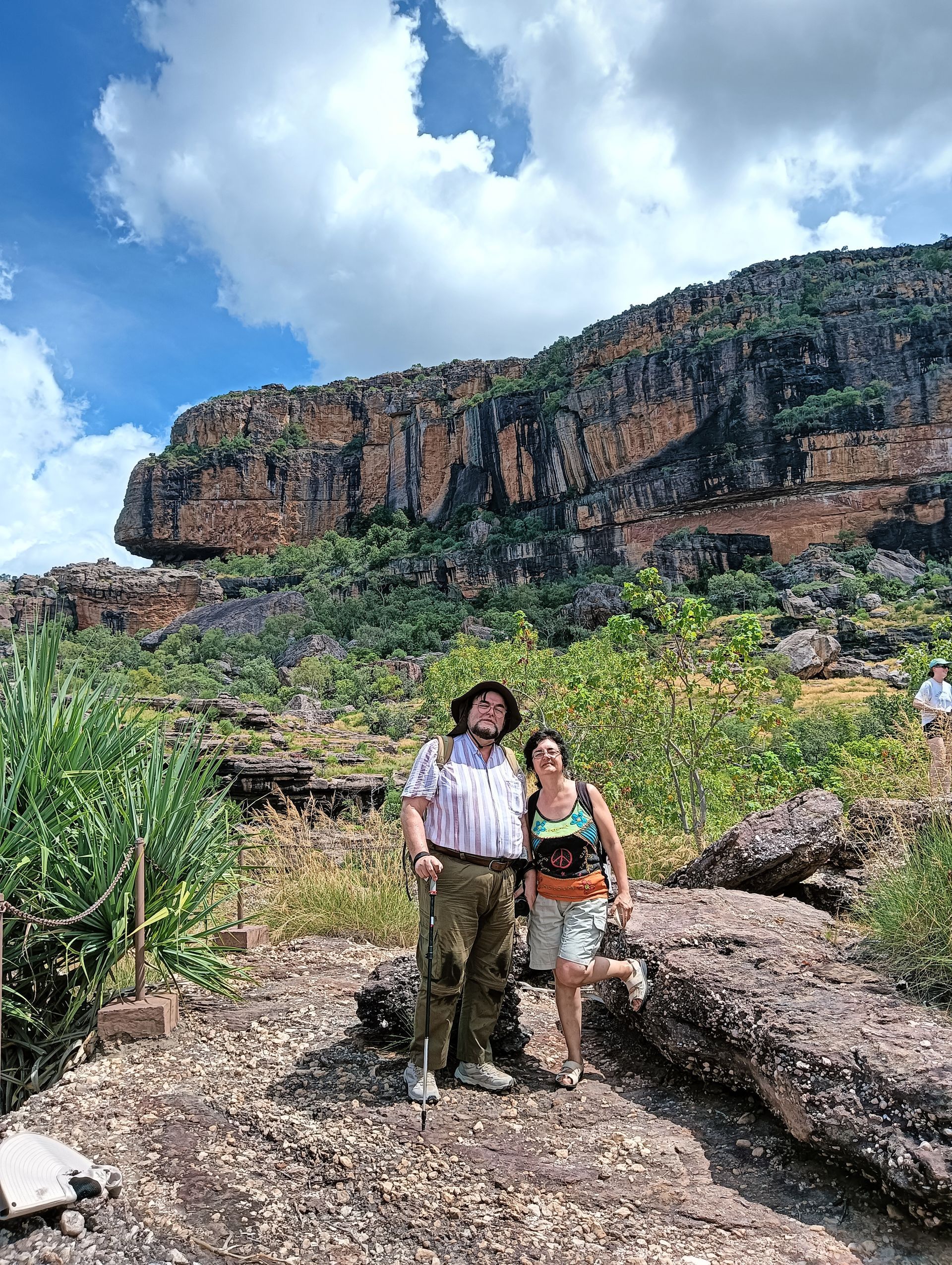 Two people stand by a rocky trail in front of a large cliff. Blue sky with clouds — NT Driver Guide - Escorted Tours & Transfers in Darwin City, NT