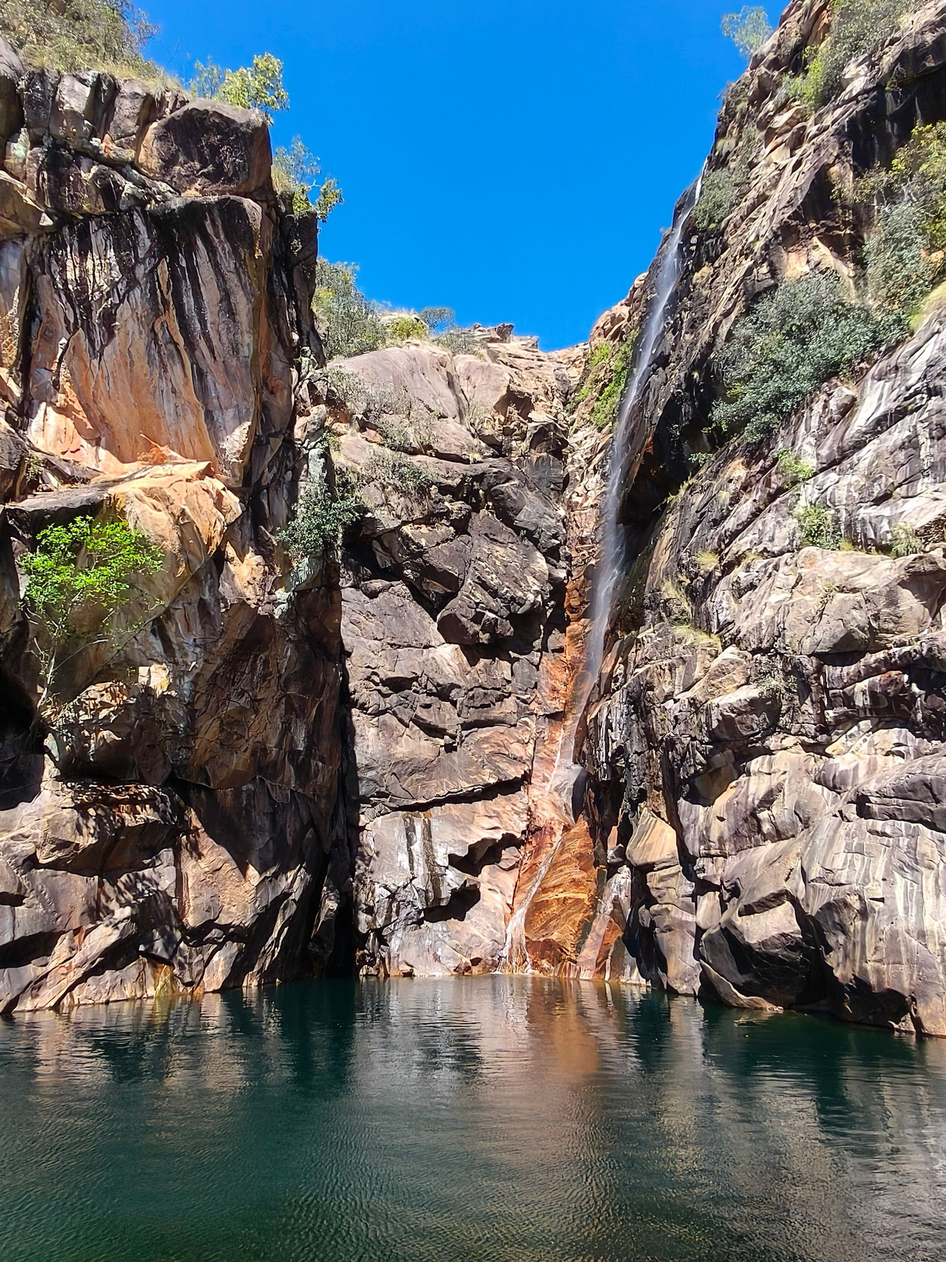 Waterfall cascading into a dark, still pool, surrounded by brown and orange rock cliffs under a blue sky — NT Driver Guide - Escorted Tours & Transfers in Darwin City, NT