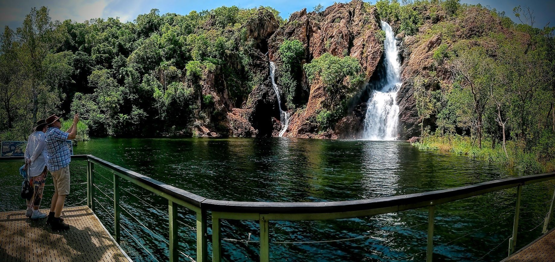 A person stands on a pier overlooking a waterfall and a green lake surrounded by trees — NT Driver Guide - Escorted Tours & Transfers in Darwin City, NT
