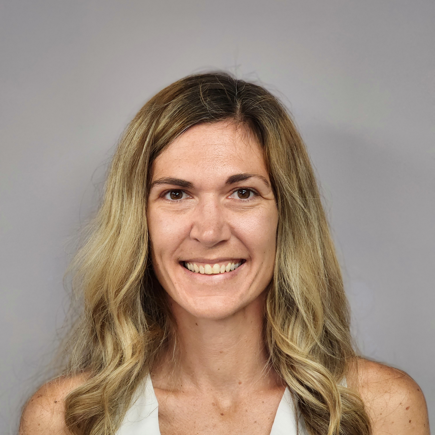 Woman with long blonde hair smiles, wearing a white top, against a gray background.
