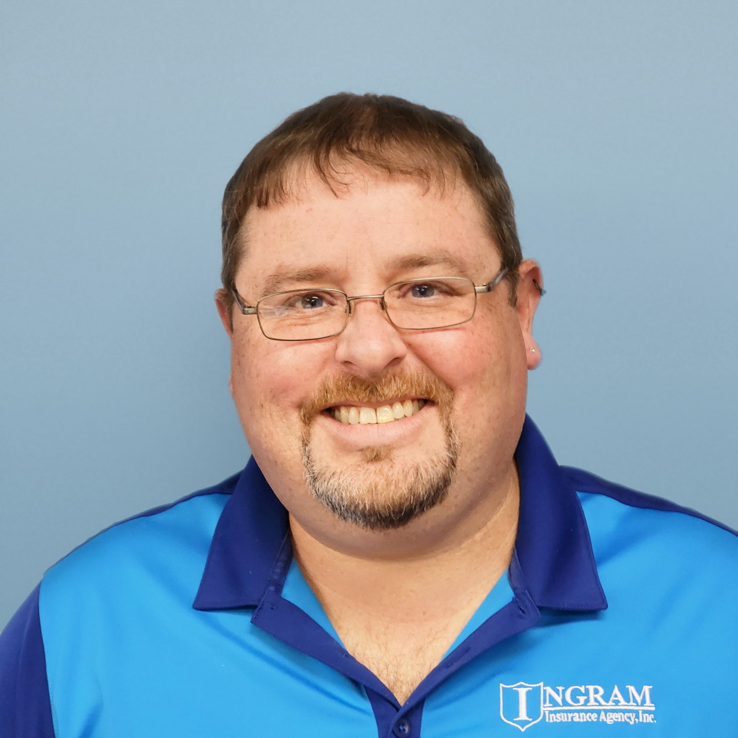 Man with glasses and goatee smiling, wearing a blue polo shirt, against a light blue background.