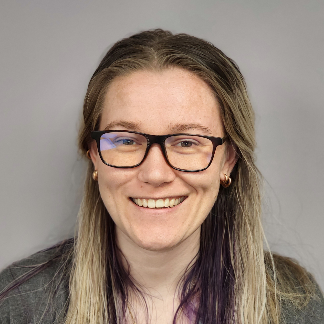 Woman with glasses, smiling, long blonde and purple hair, wearing a gray shirt, in front of a gray wall.