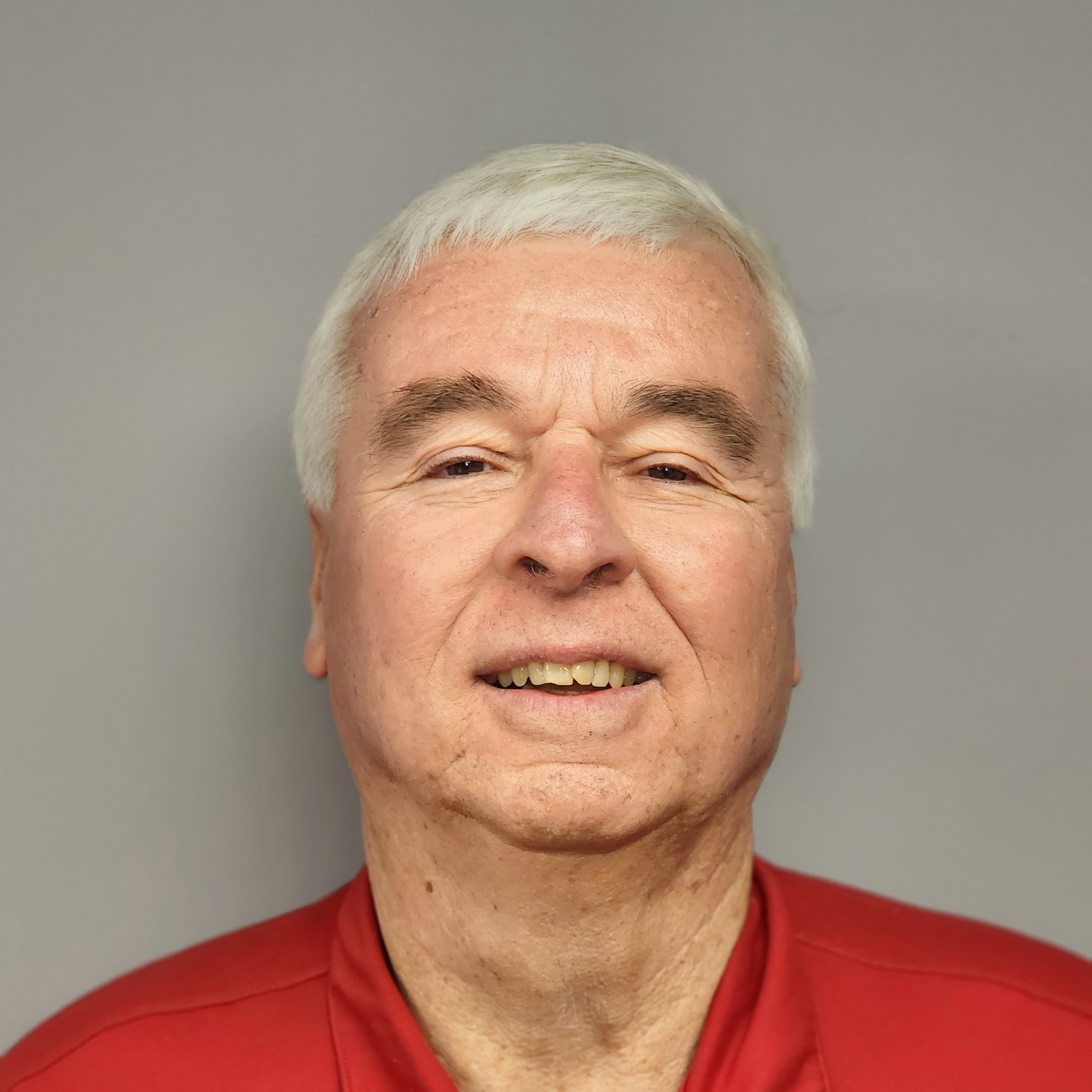 Man with gray hair and red shirt smiles at the camera against a gray background.