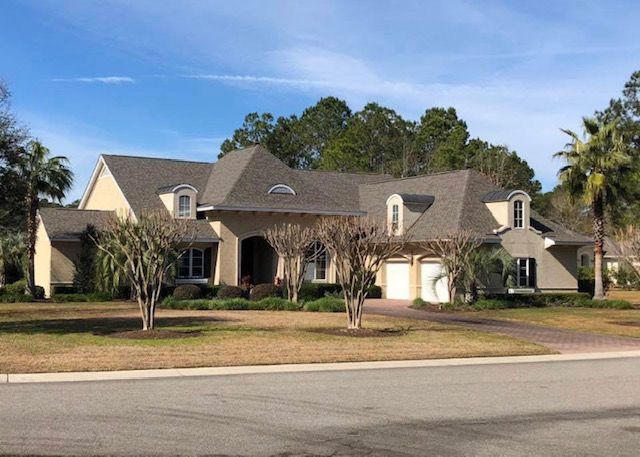 Large beige suburban house with a gray roof, white garage, and palm trees beside a street