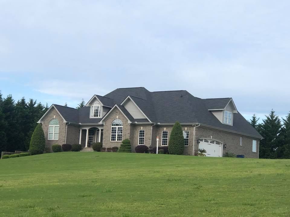 Large brick suburban house with a dark roof on a grassy hill under a cloudy sky