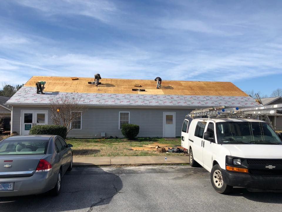 House roof under construction with exposed plywood, parked cars in front, and a clear blue sky.