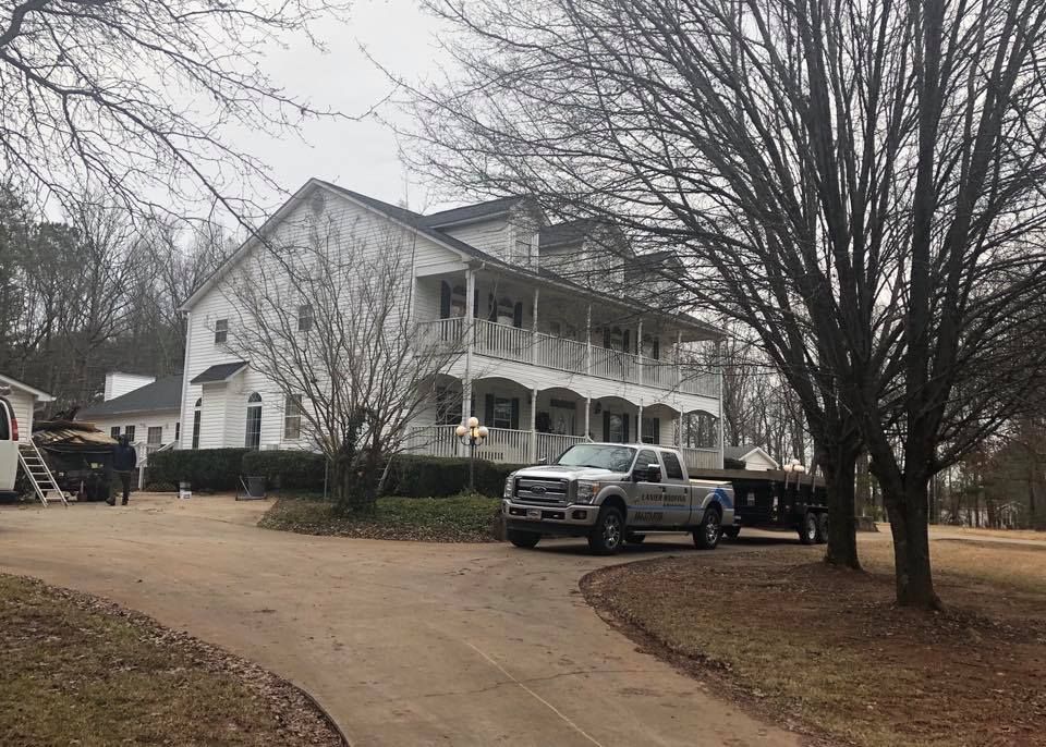 White two-story house with wraparound porch and parked pickup trucks on a curved driveway under bare trees