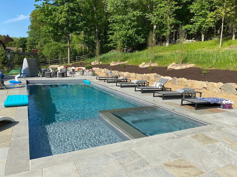 A rectangular pool with a built-in spa, surrounded by lounge chairs on a stone patio with a grassy hillside background.
