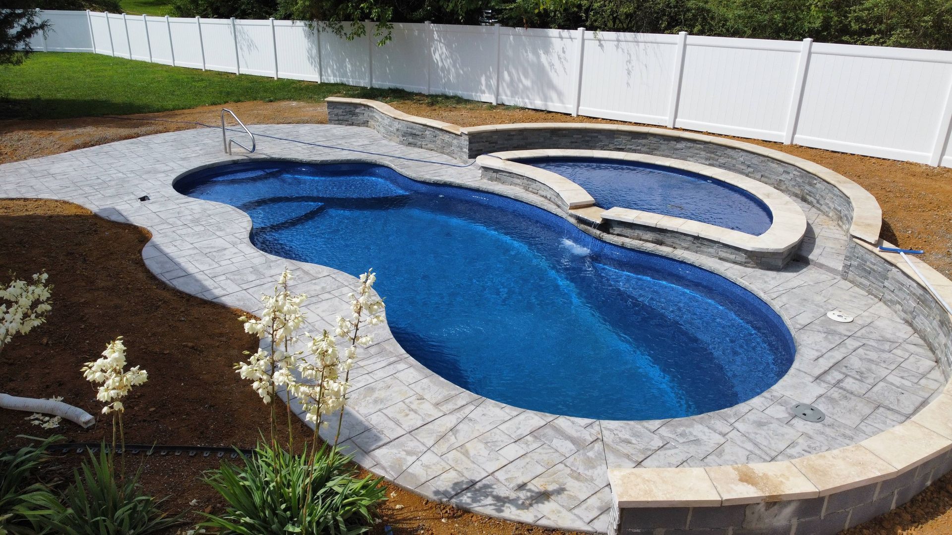 A backyard swimming pool with a connected hot tub, surrounded by gray stone pavers and a white vinyl fence.