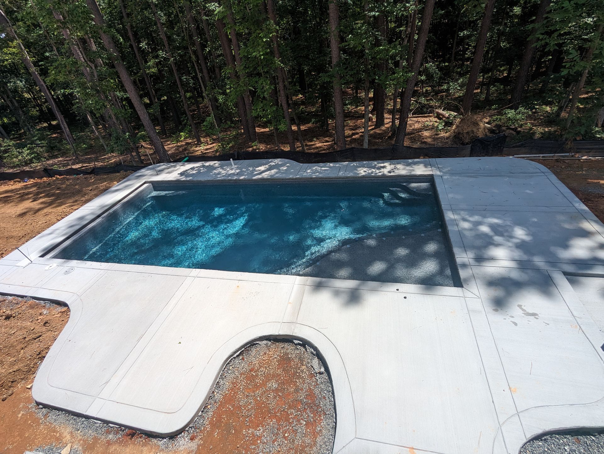 A rectangular blue swimming pool surrounded by a newly poured, light gray concrete deck in a wooded backyard setting.