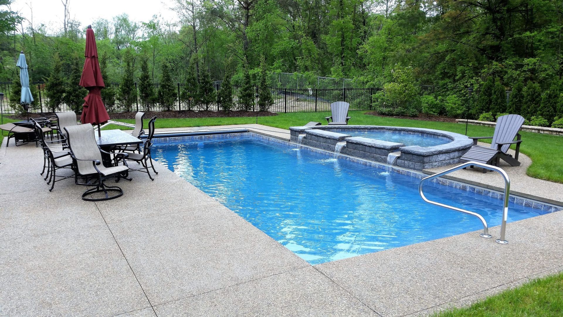 A rectangular blue swimming pool with an attached raised hot tub, surrounded by a light stone patio and green trees.