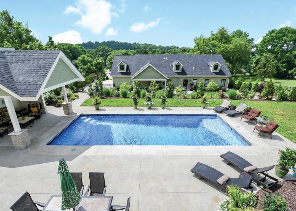 A rectangular swimming pool set in a backyard patio with lounge chairs, a covered seating area, and a large house beyond.