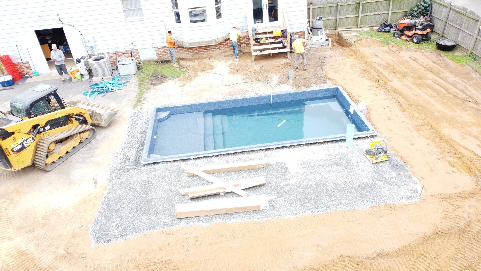 An aerial view of a rectangular, dark blue swimming pool under construction in a residential backyard with heavy machinery.
