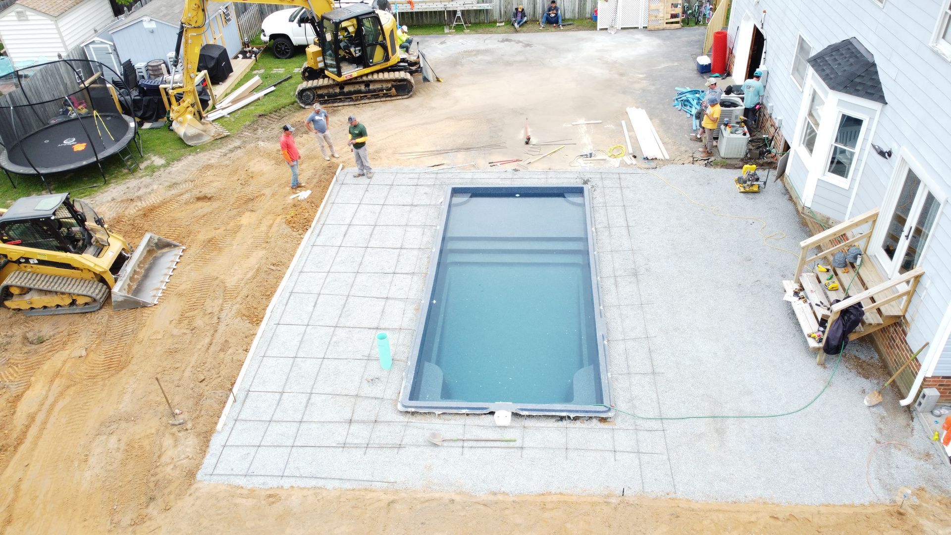 An aerial view shows a rectangular pool installed in a backyard construction site with workers and heavy machinery nearby.