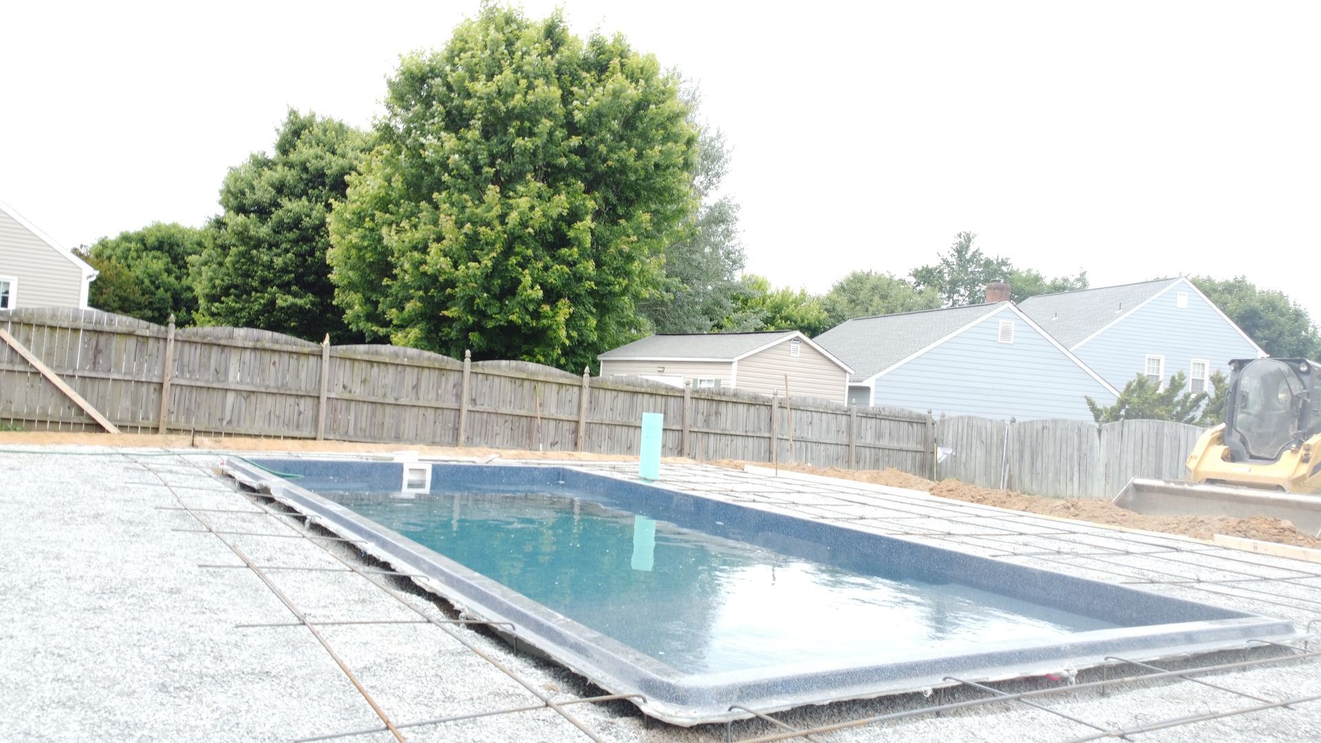 A rectangular pool sits in a gravel-covered backyard with a wooden fence and houses in the background.