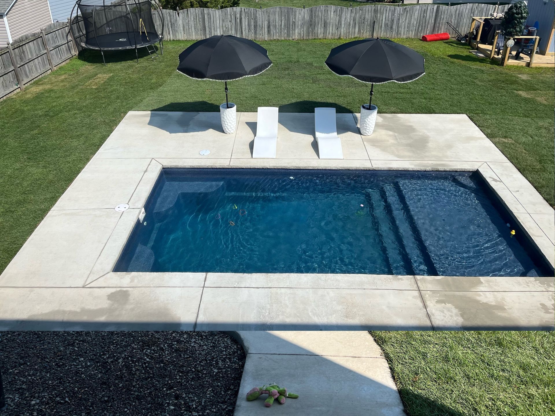 A rectangular inground pool with two white lounge chairs and black umbrellas on a concrete patio in a grassy backyard.