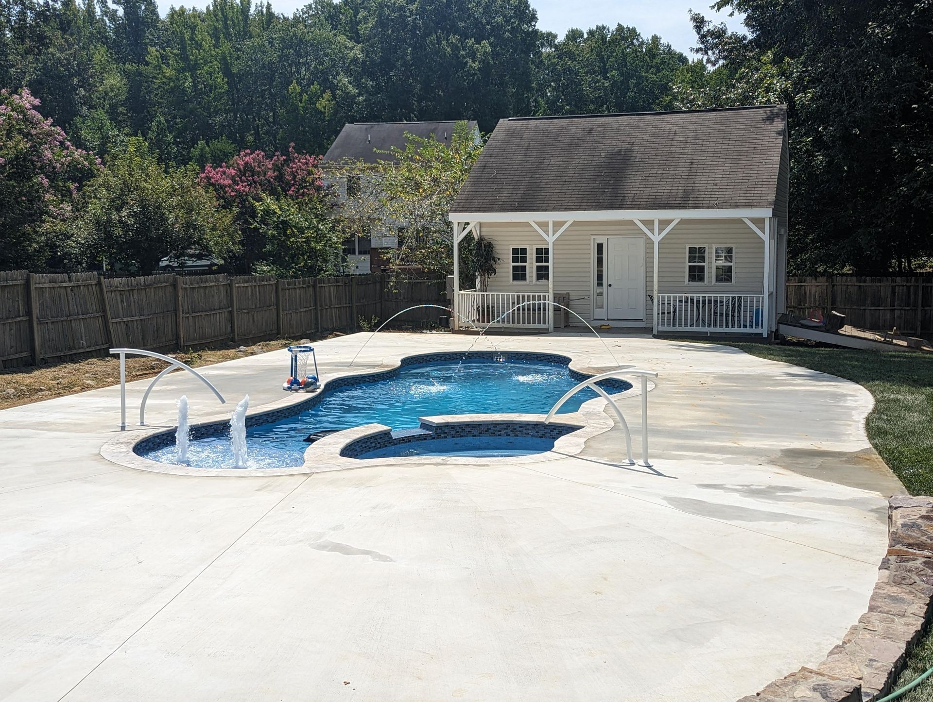 A pool with water fountains and an attached spa on a large concrete patio in front of a small house with a porch.