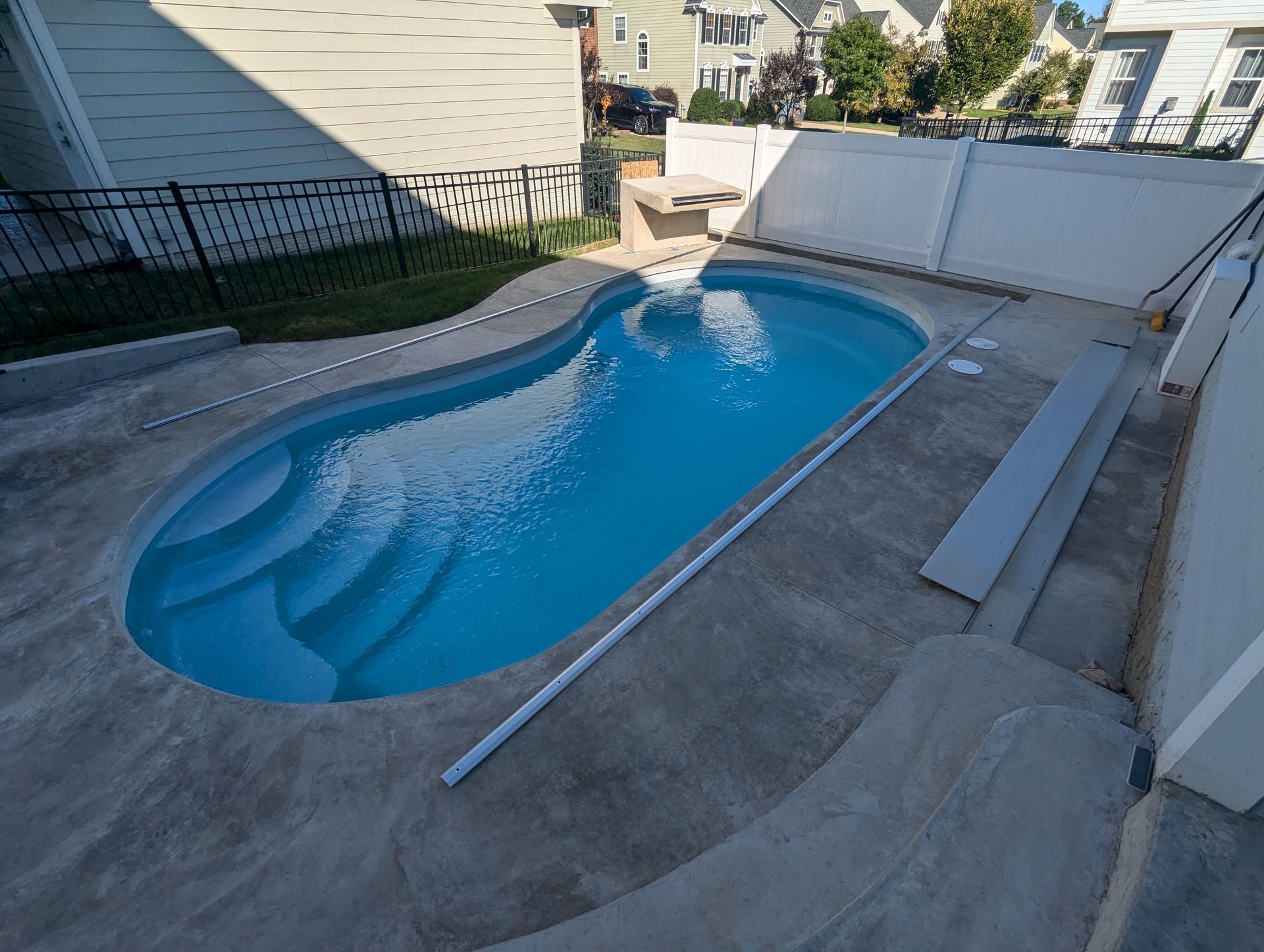 A backyard swimming pool with blue water and built-in steps, surrounded by a gray concrete patio and white vinyl fence.