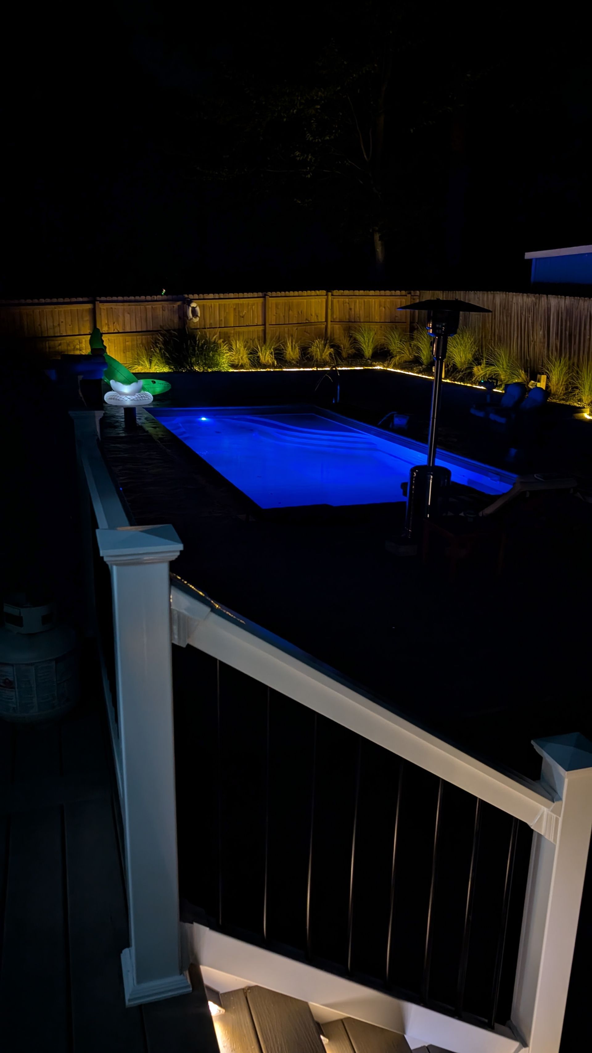 A backyard deck at night with a glowing blue rectangular pool and warm landscape lighting along a wooden fence.