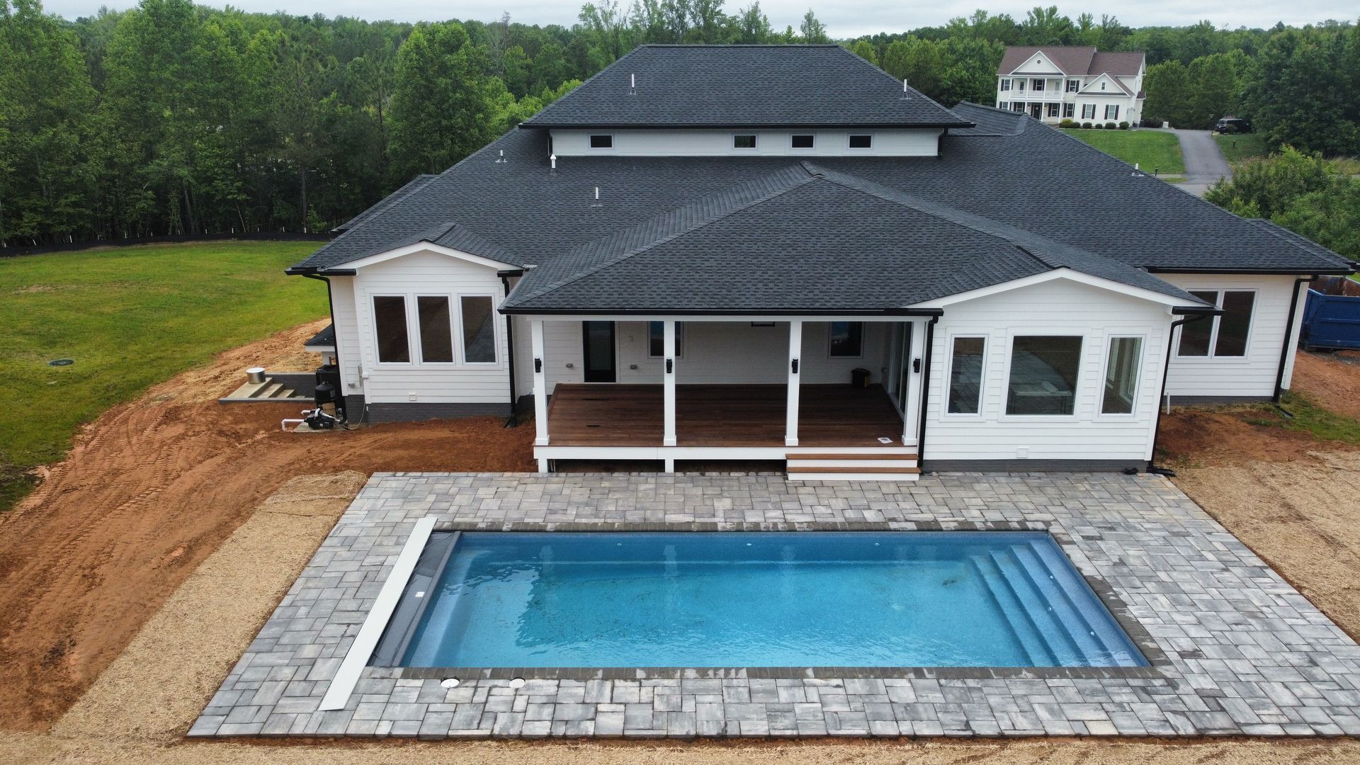 An aerial view of a white, multi-roofed home with a large backyard pool surrounded by a stone patio and bare earth.