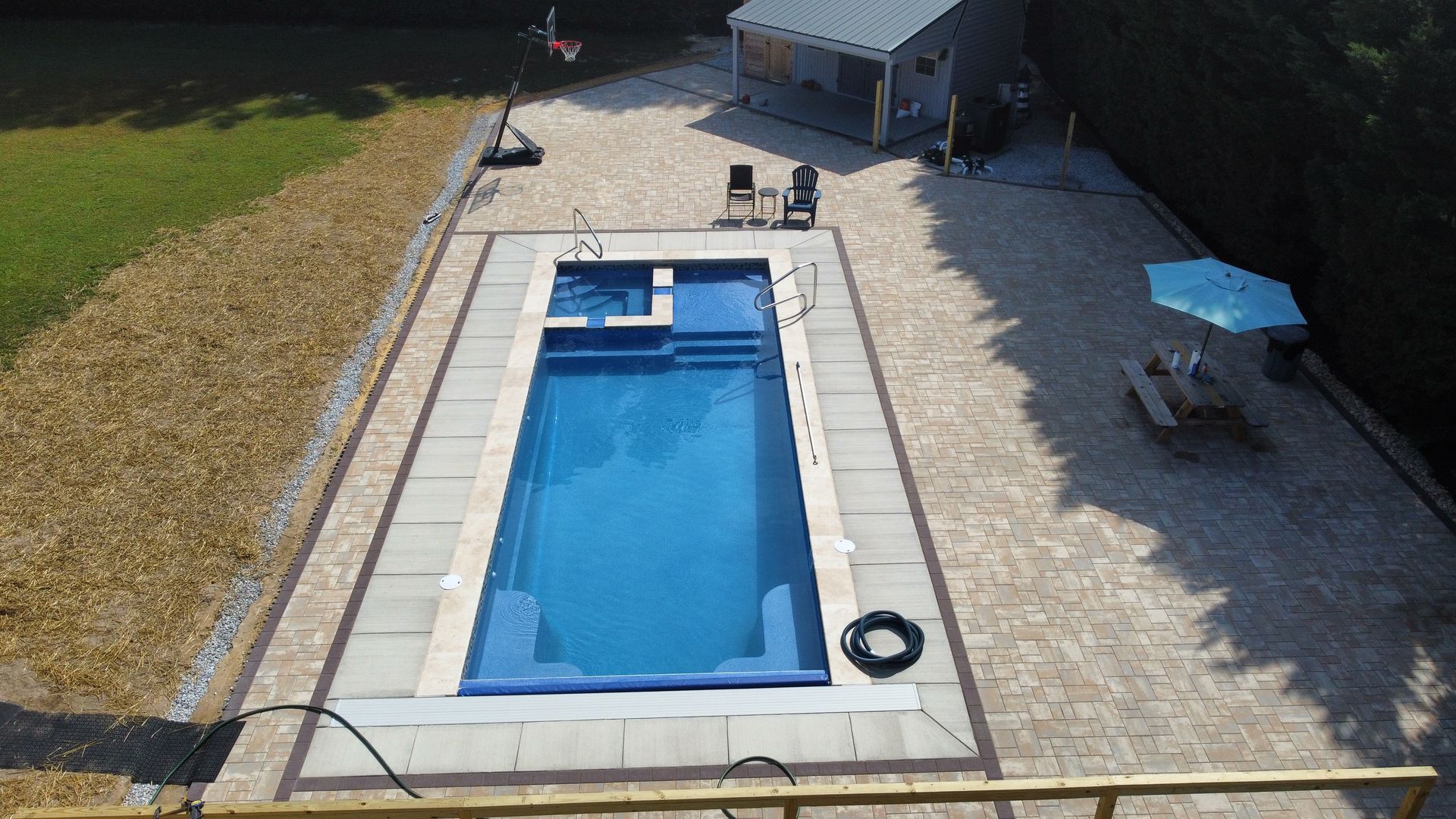 A rectangular inground pool with light-colored pavers, a small cabana, a picnic table with a blue umbrella, and grass nearby.